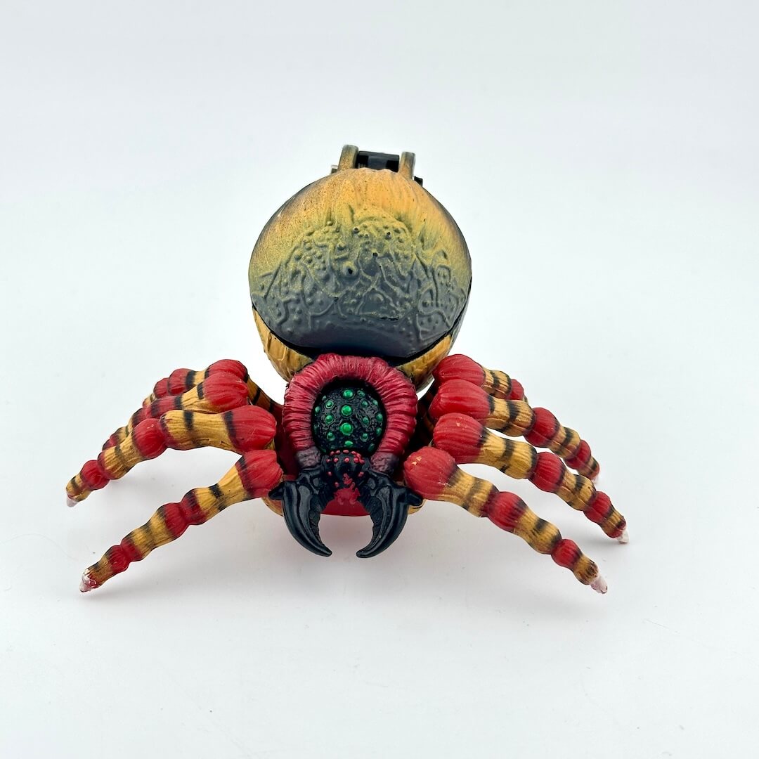 A spider action figure with a textured gold and black abdomen, red and yellow banded legs, and a central green eye.