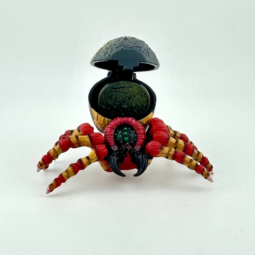 A spider action figure with open back to show coin detail, spider has red and yellow legs and gray body. 