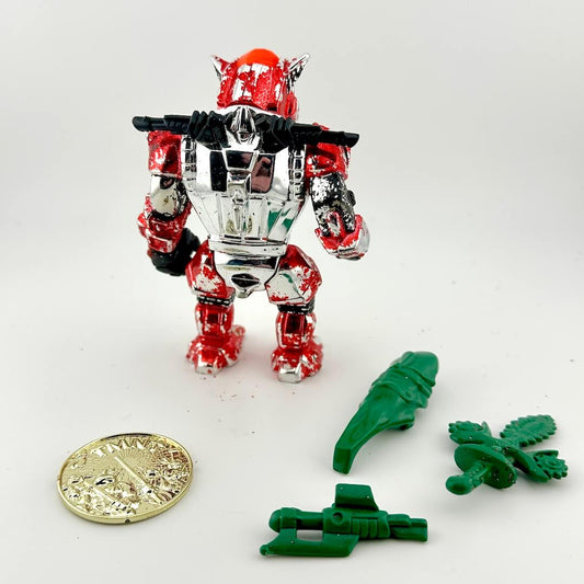 Toy figure with red, silver, and black color scheme on a white background with green accessories and a coin for scale.