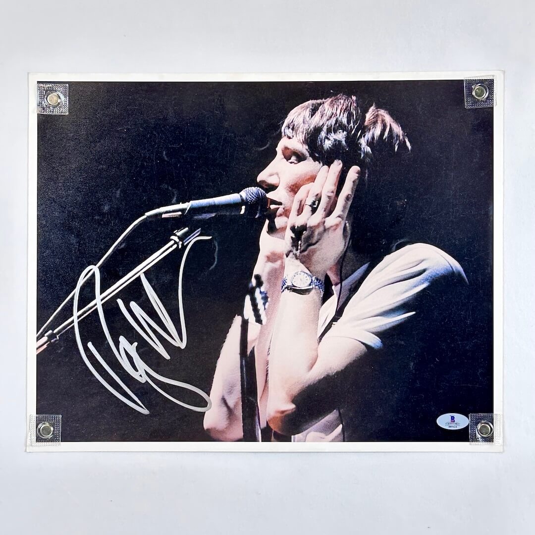 Person singing into a microphone with a dark background, signature in silver at left.