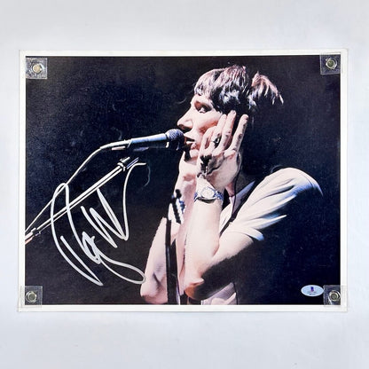 Person singing into a microphone with a dark background, signature in silver at left.