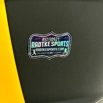 A close-up of a black helmet with a yellow stripe on the left side, featuring a custom shield-shaped sticker for 'Radtke Sports.'