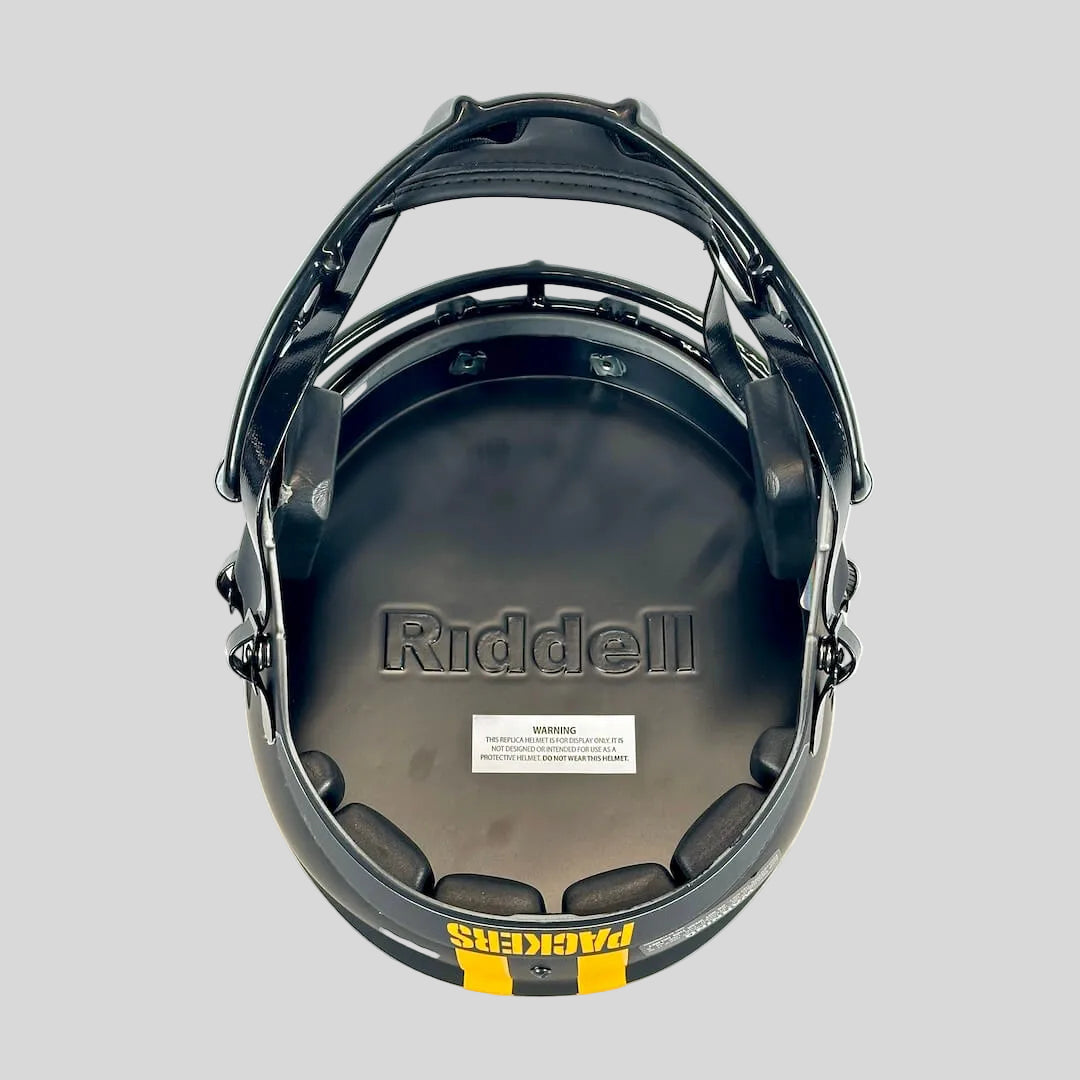Black Riddell football helmet underside with padded interior, visible Riddell branding, a small warning label, and a yellow/black Packers logo strip at the bottom.
