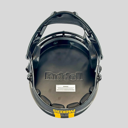 Black Riddell football helmet underside with padded interior, visible Riddell branding, a small warning label, and a yellow/black Packers logo strip at the bottom.