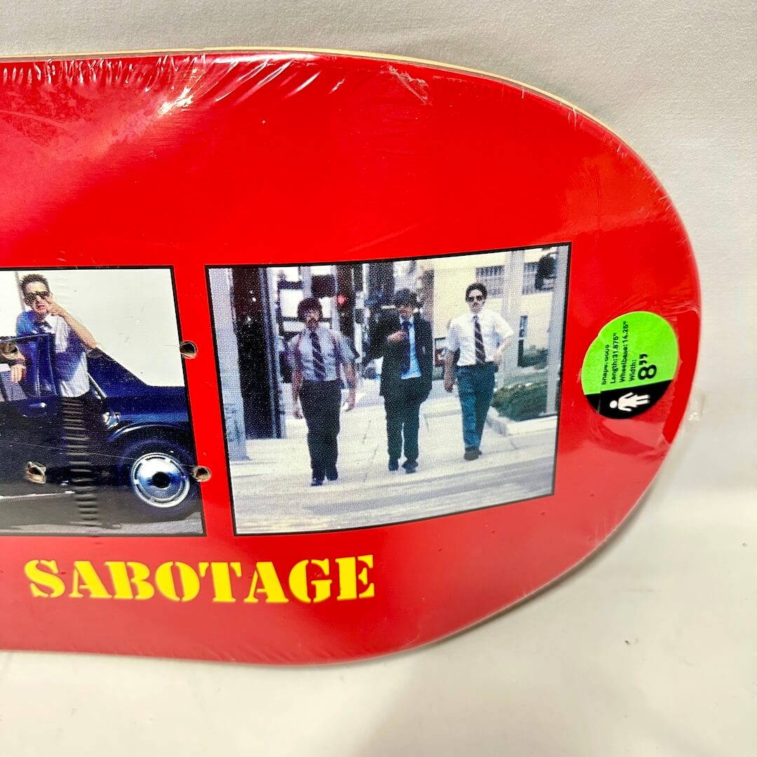 A red skateboard deck wrapped in plastic, featuring two photos and the word 'SABOTAGE' in yellow text at the bottom. 