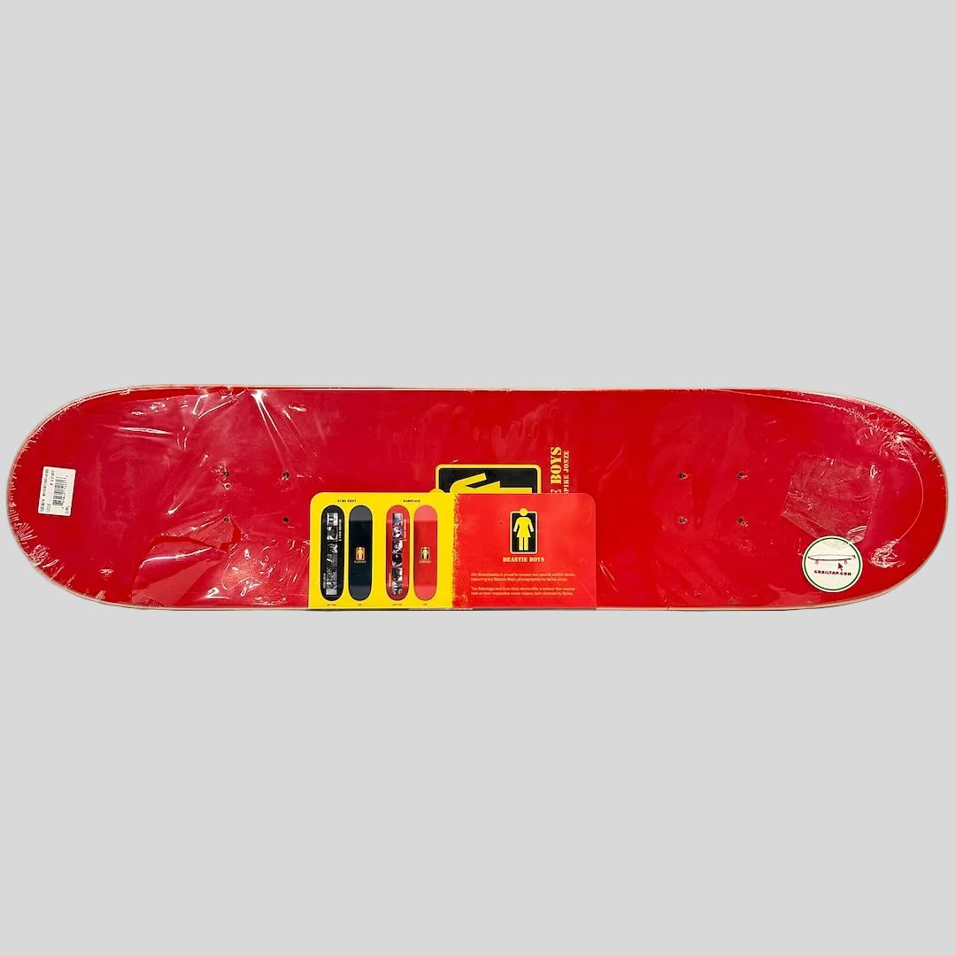 A red skateboard deck shown from the underside, still wrapped in protective plastic, with a yellow and red retail card. 