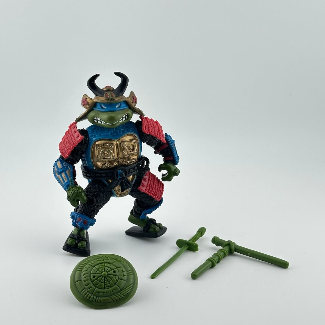 Anthropomorphized turtle wearing blue, gold and red samurai armor, in blue mask, pictured with green accessories.