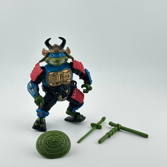 Anthropomorphized turtle wearing blue, gold and red samurai armor, in blue mask, pictured with green accessories.