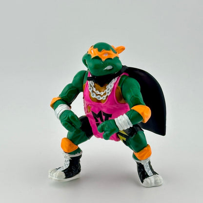 Anthropomorphized turtle wearing pink jersey, orange protective gear, black cape, & white jewelry. Front.