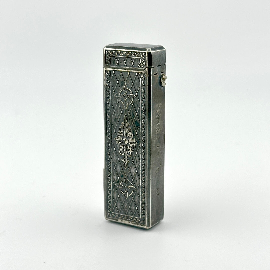 A vintage silver pocket lighter with ornate engraved floral and geometric patterns on its rectangular body.