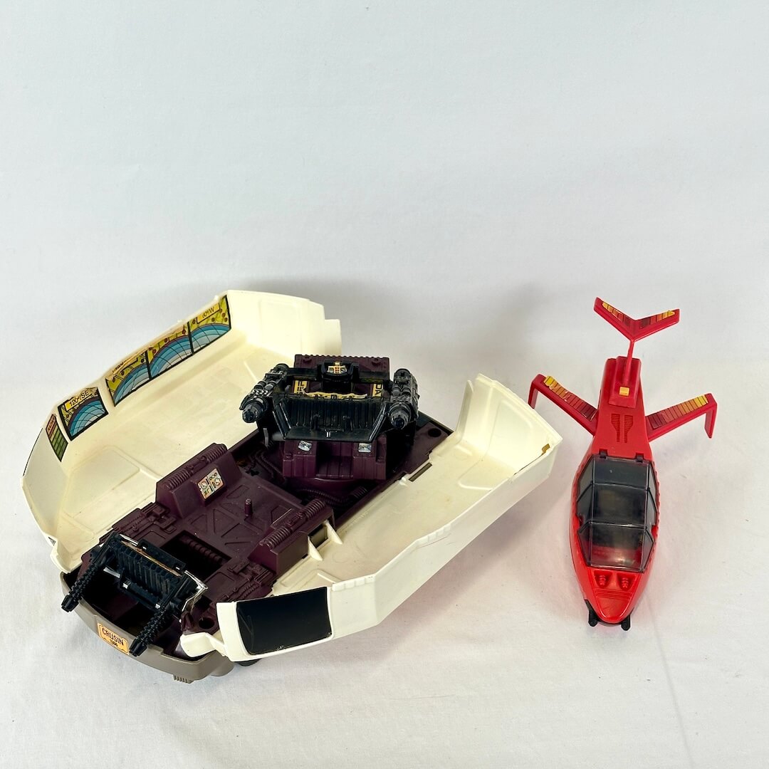 A vintage toy set featuring a red helicopter and a beige and dark purple vehicle inside its open cream-colored shell.