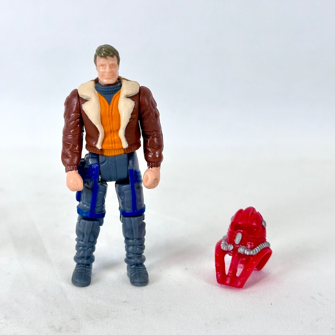 A vintage action figure of a male pilot standing next to a small red helmet. 
