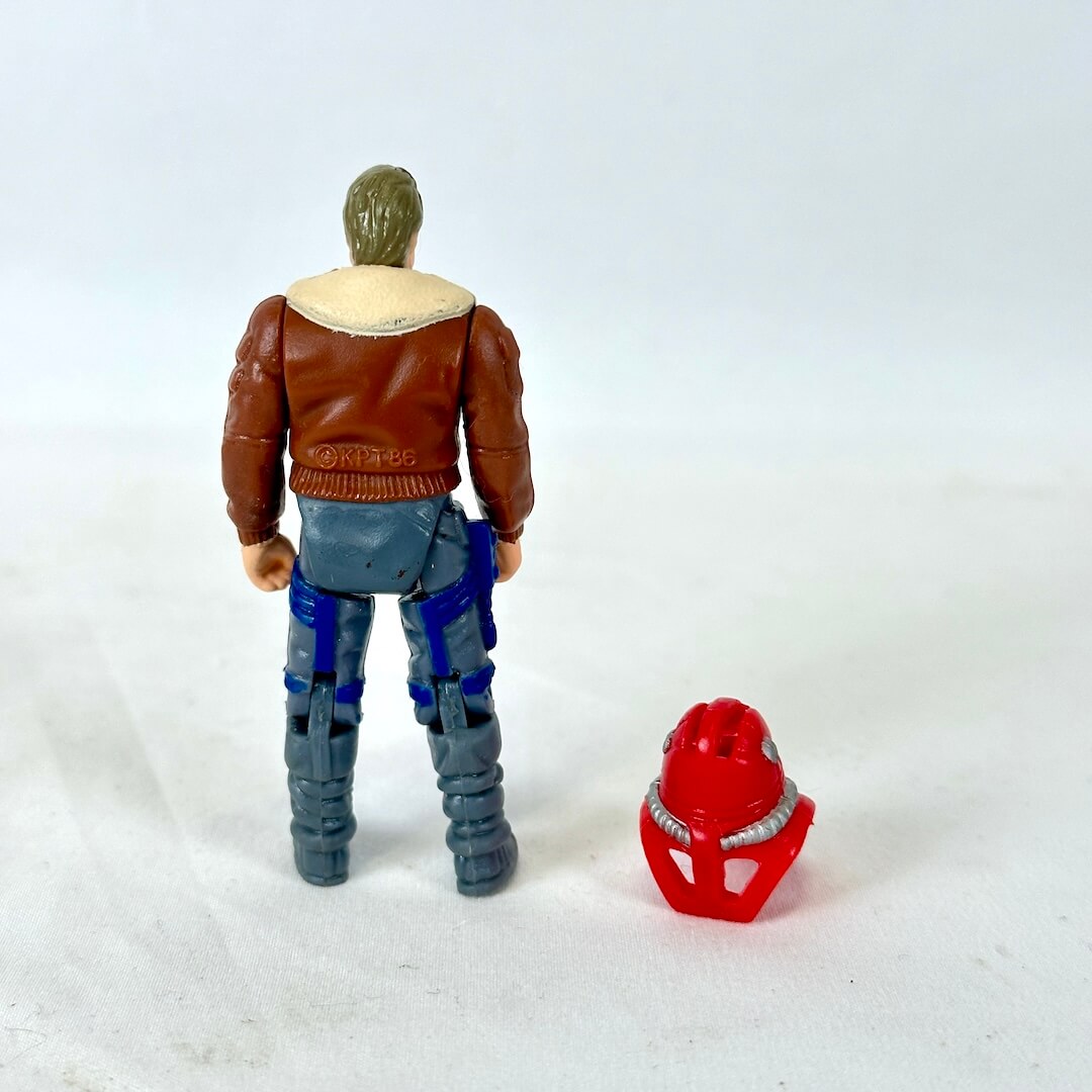 Action figure shown from the back, wearing a brown jacket, blue pants, and gray boots, standing next to a small red helmet. 