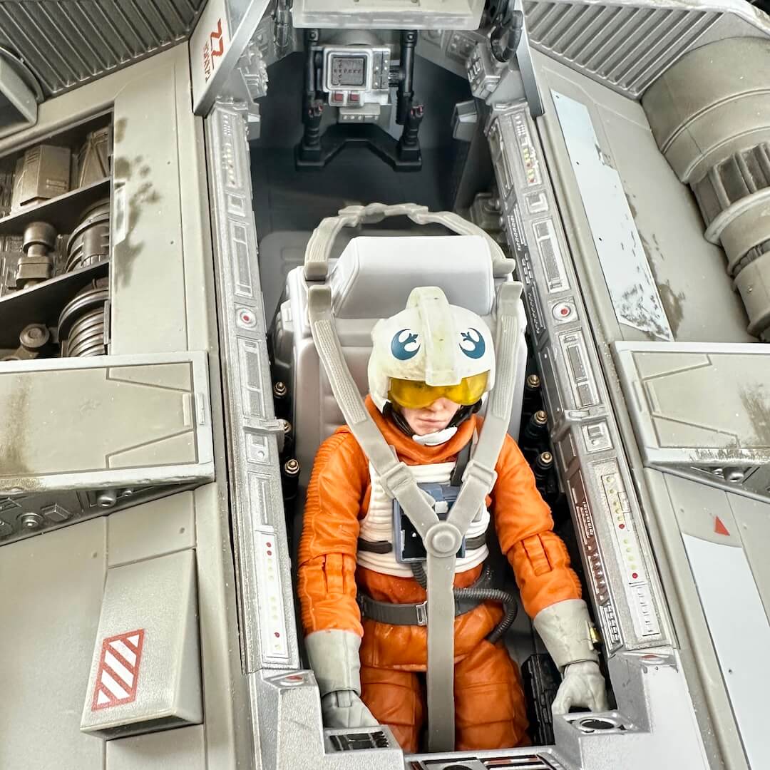 A person wearing an orange spacesuit with a white helmet and yellow visor seated inside a gray sci-fi spacecraft cockpit.