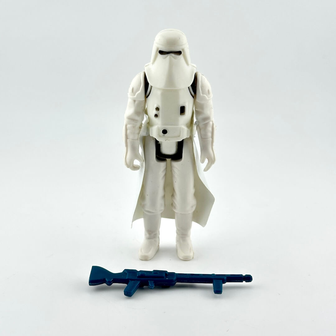 A white action figure standing upright on a light gray background, with a blue rifle accessory placed in front of it.