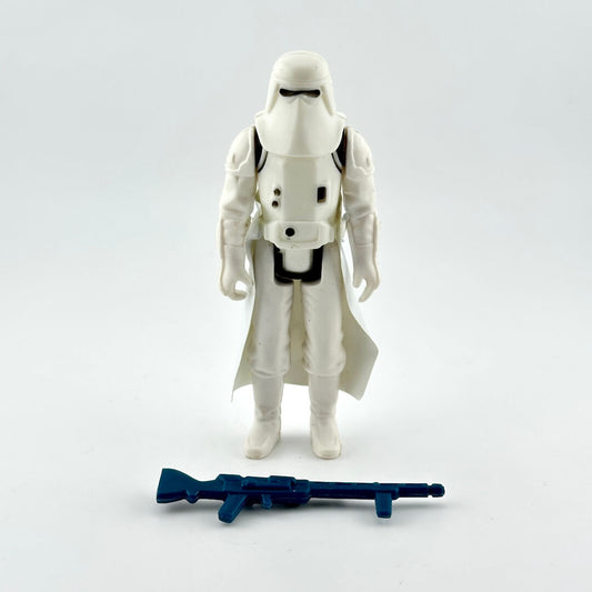 A white action figure standing upright on a light gray background, with a blue rifle accessory placed in front of it.