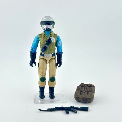 Action figure in a futuristic outfit with a helmet, vest, and pants, standing next to a brown sandbag and a weapon on a white background.