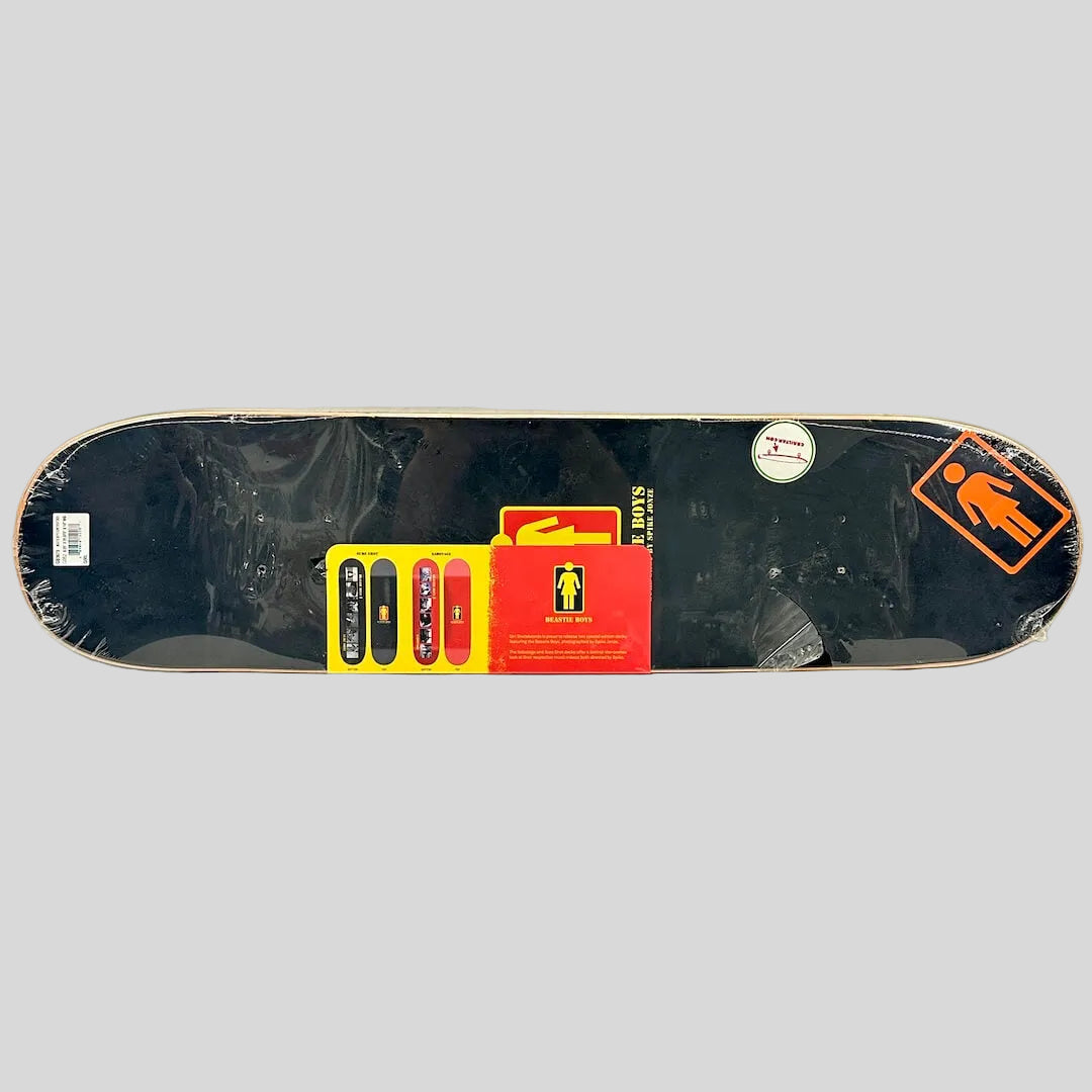 A black skateboard deck with stickers and protective plastic wrap, displayed horizontally.