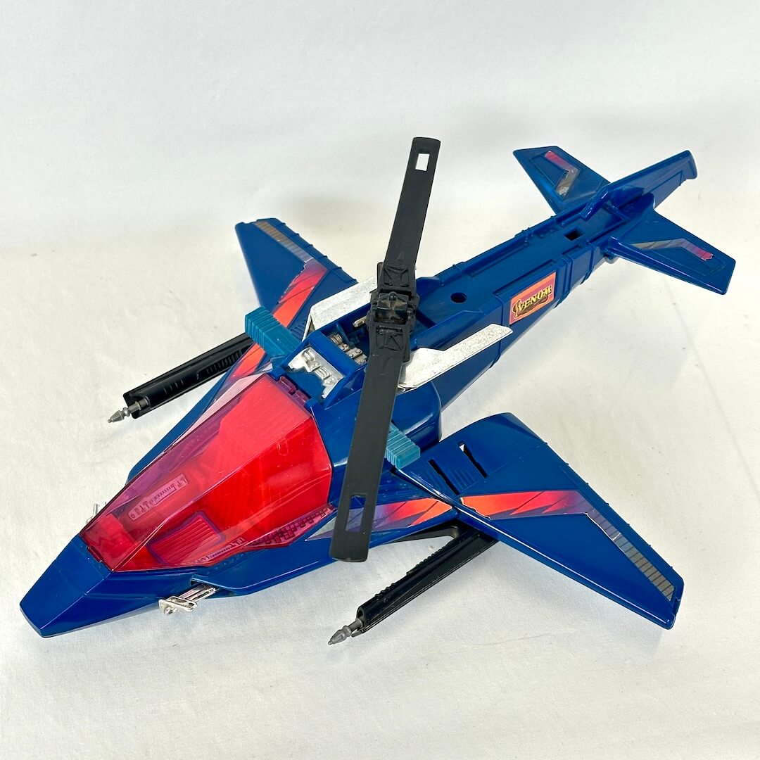 A blue and red toy jet helicopter with black rotor blades, photographed on a white fabric background.