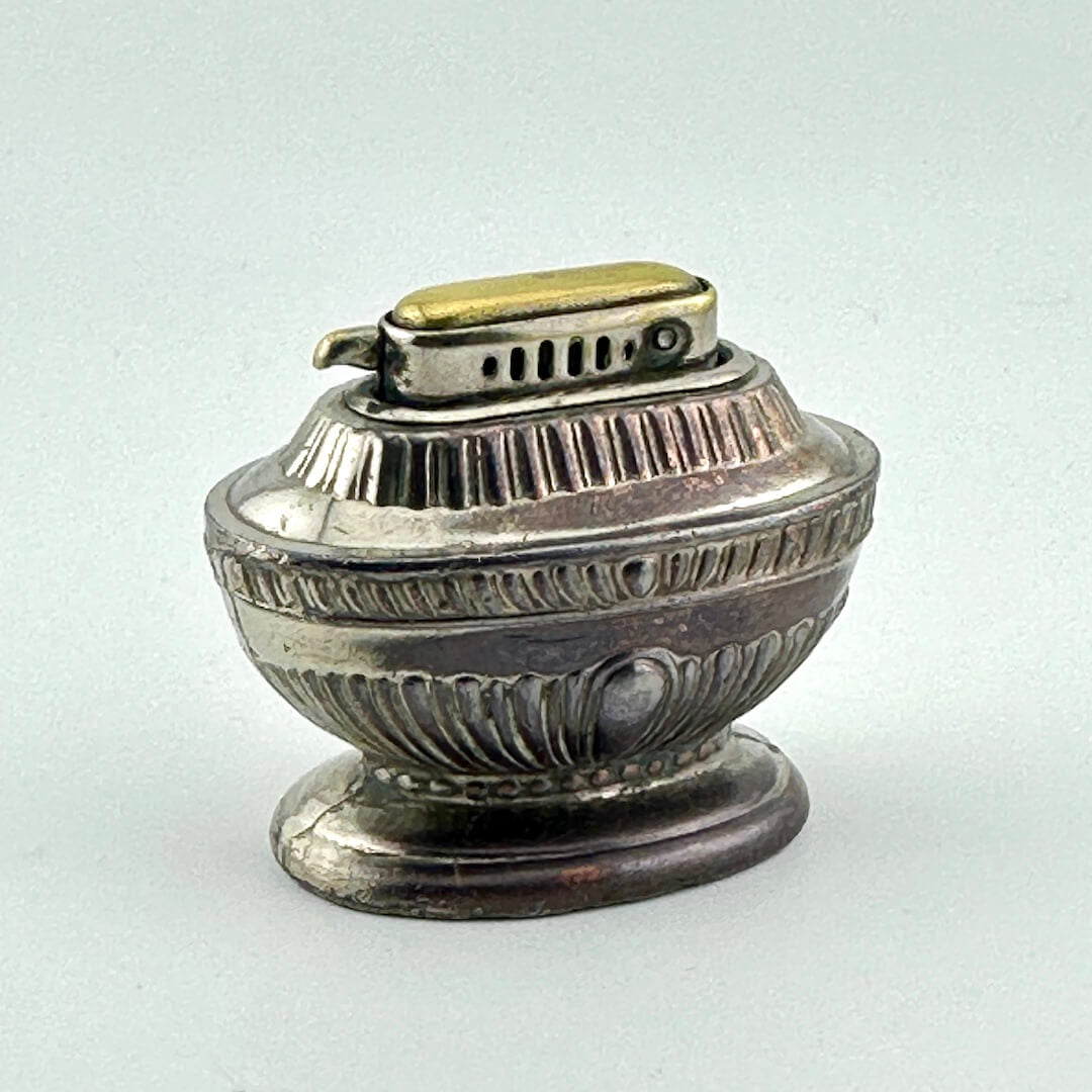 A vintage silver-toned metal lighter with a decorative ribbed body and  brass on top, set against a plain light gray background.