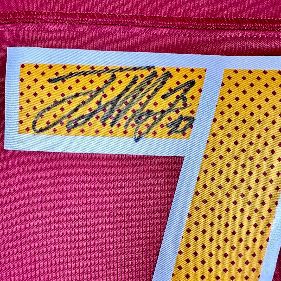 A close-up of a red textured fabric, featuring a white-bordered patch with a yellow background, and signature. 