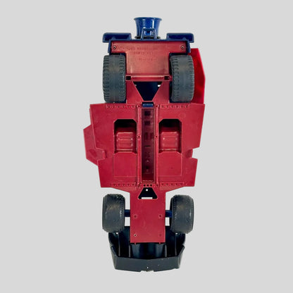 A vintage red toy truck shown upside down, revealing its black wheels and blue top mechanism.