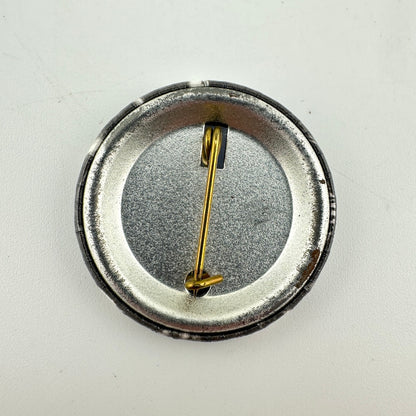 The back of a round pinback button badge with a black outer rim, and a gold-tone safety pin clasp.