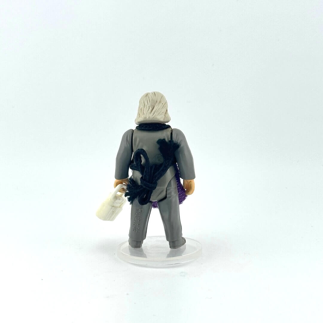 Alien action figure in gray outfit and purple apron holding white briefcase against white background.