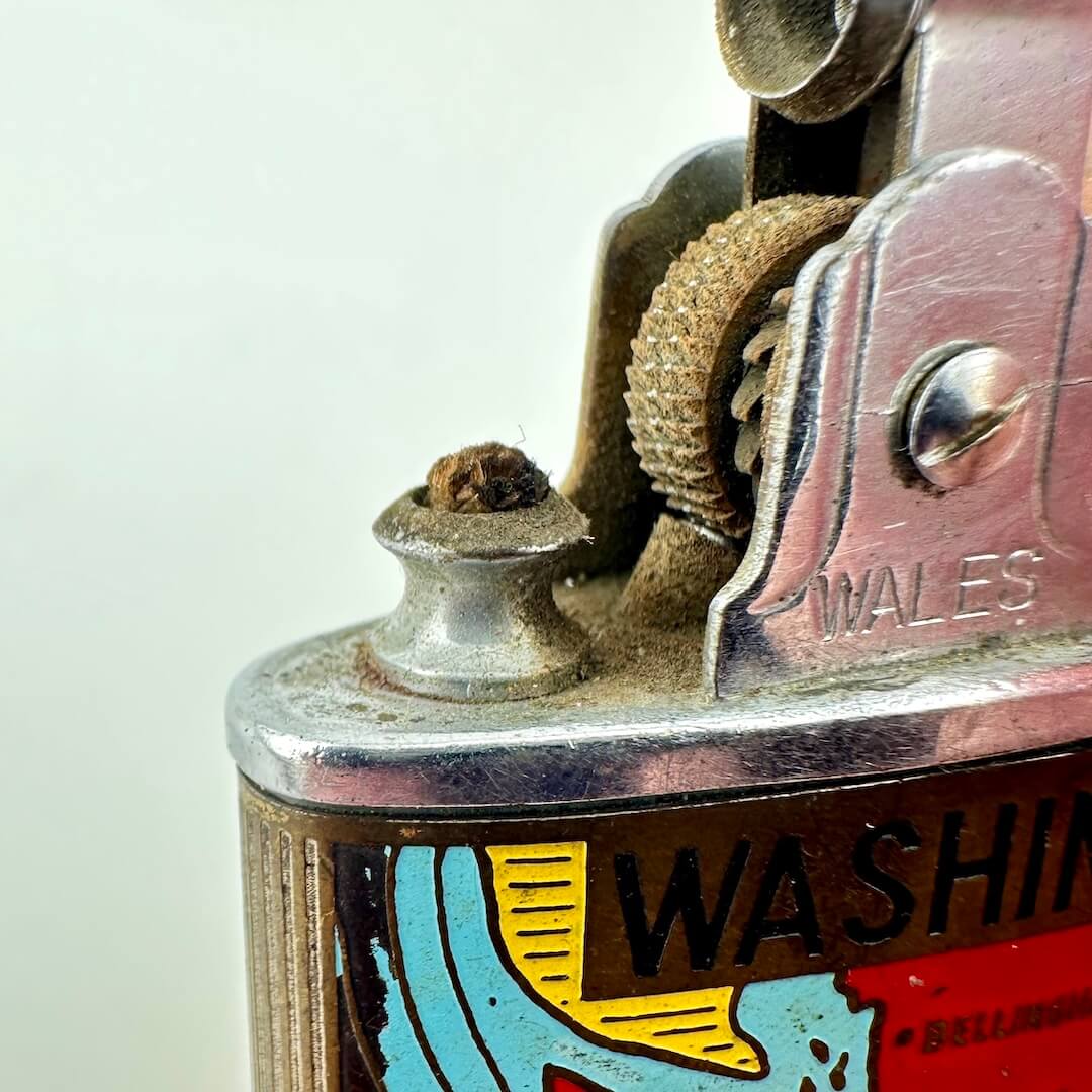 Close-up of a vintage metal lighter with a colorful label reading WASHINGTON, and a small brown tip on the wick.