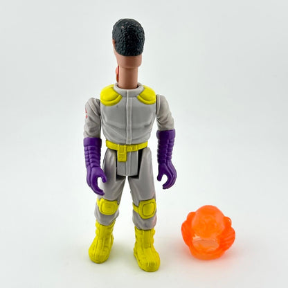 Toy figure in gray and yellow outfit with purple gloves and black hair, standing next to an orange object on a white background.