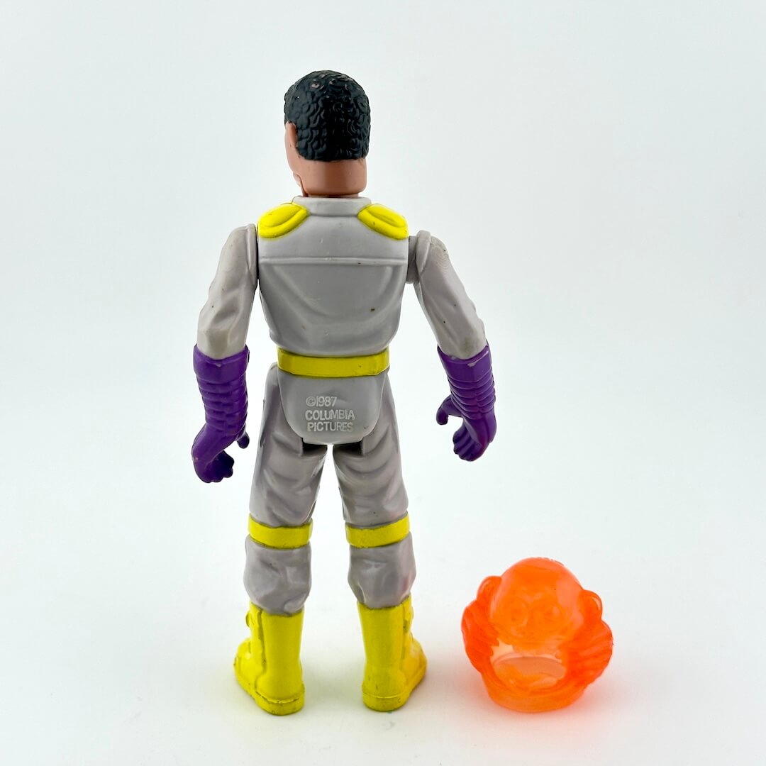 Toy figure in a gray suit with purple gloves and yellow boots, standing next to an orange object on a white background.