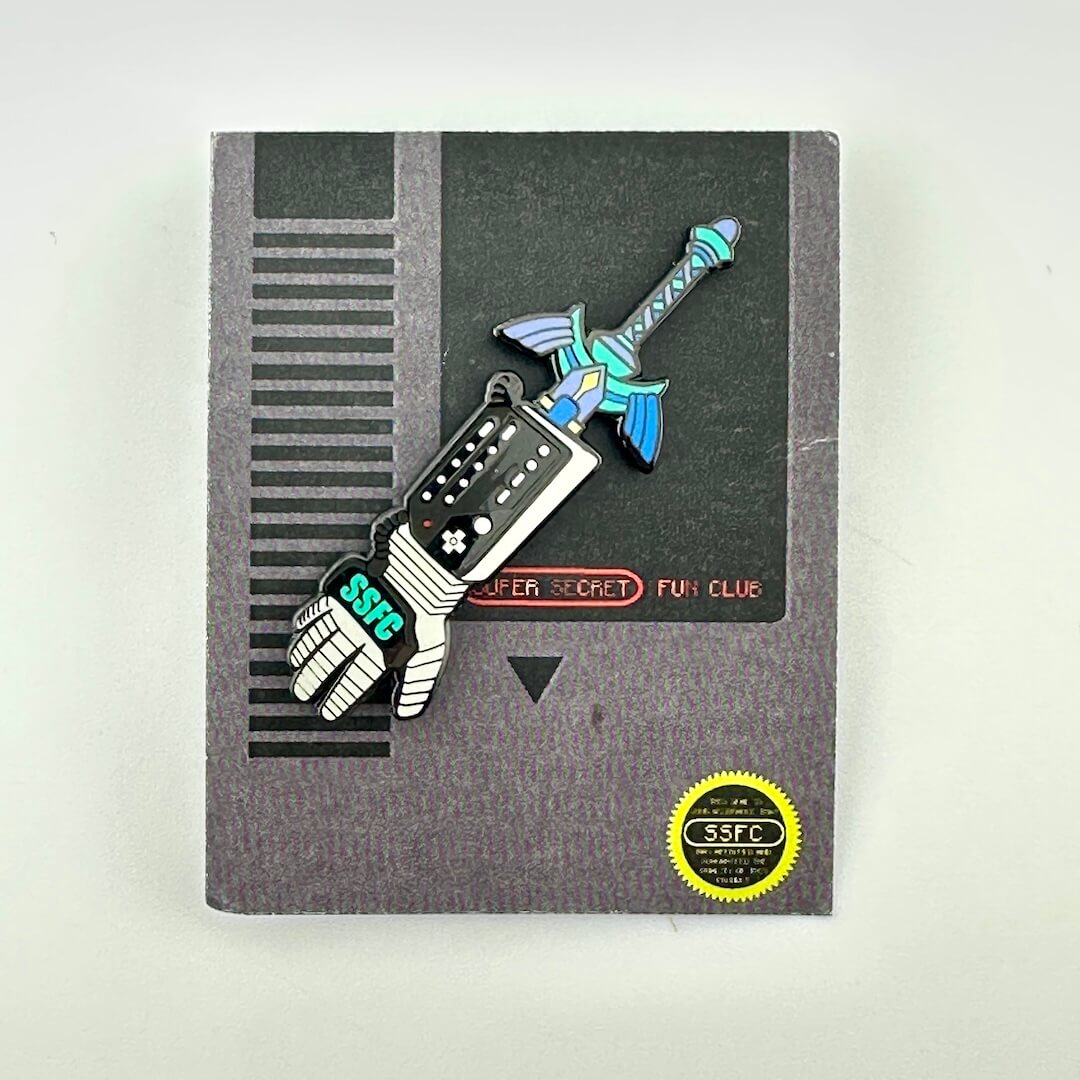 Enamel pin of sword sticking out of the top of futuristic glove with buttons, on dark card. 